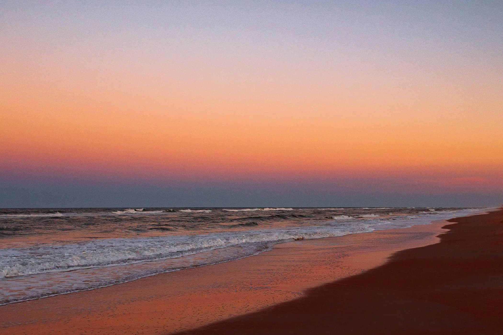 Quiet beach shoreline at sunset with soft waves and a pastel-colored sky.